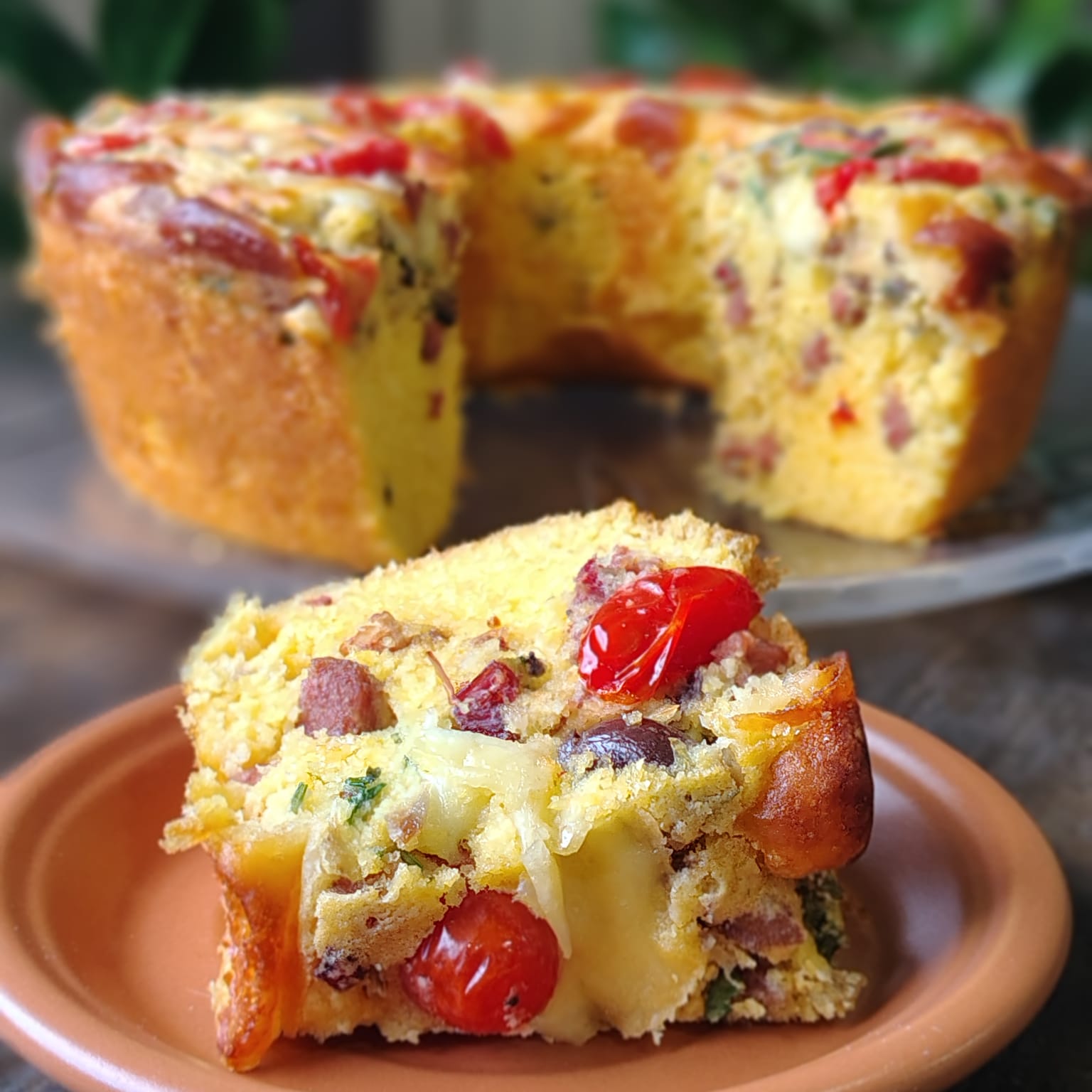 Fatia de bolo salgado de milho com calabresa, bacon, azeitona e tomate, servida em prato de barro